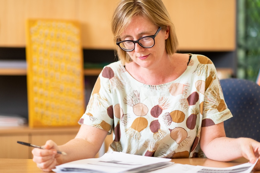 A woman wearing glasses sits at a desk peering at paper and holding a pen. 