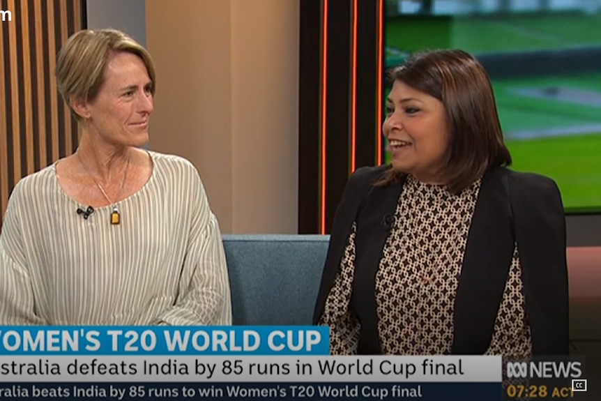 Rica sits with another guest on a blue sofa in a TV studio with a backdrop showing the cricket on TV screens