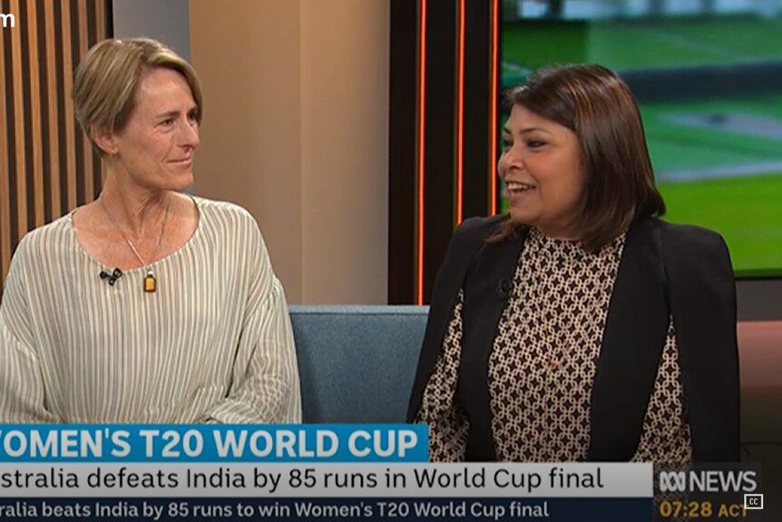 Rica sits with another guest on a blue sofa in a TV studio with a backdrop showing the cricket on TV screens
