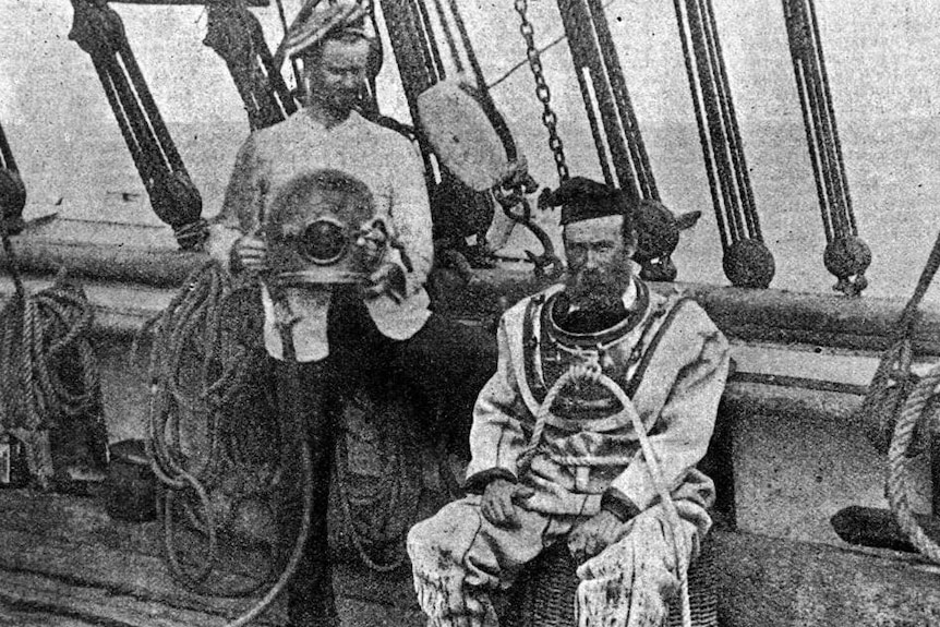 black and white photo of rigging boat with vintage diver sitting on basket and crew member on left with helmet.