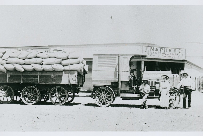 An old brown photograph of a truck hauling a carriage with a woman and a man standing in front of it.