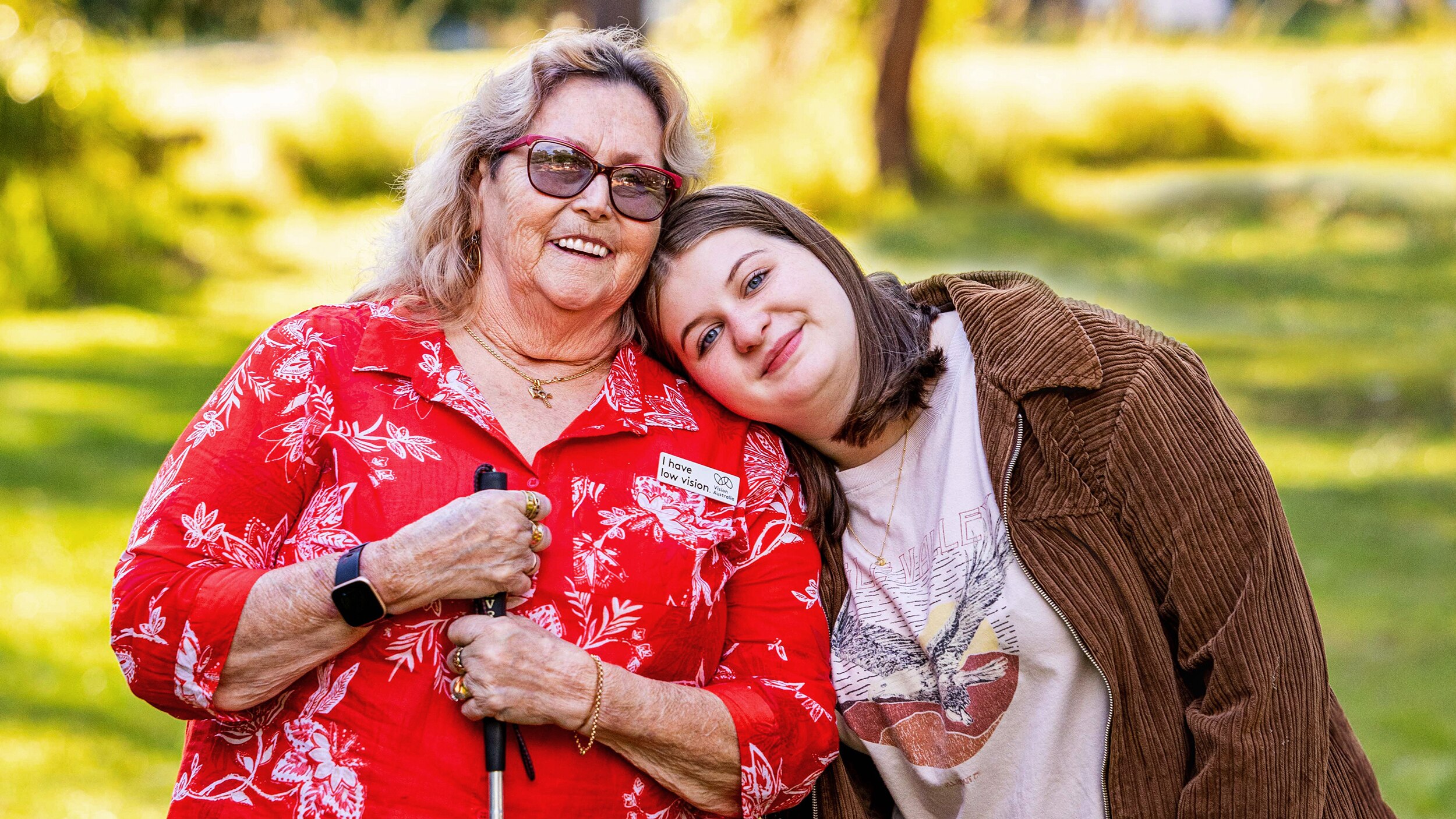A teenager rests her head on the shoulder of an older woman who’s wearing tinted glasses and holding a white cane for low vision