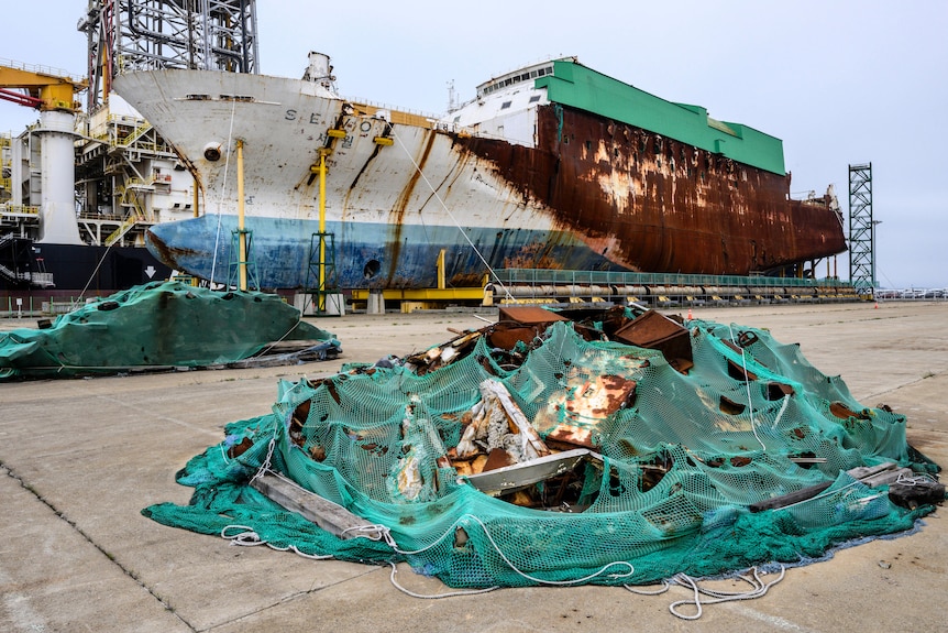 A dilapodated wrecked ship sits on a dry dock near green marine netting. 