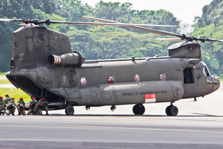 A large grey Chinook helicopter with Singapore flag on the side is grounded, with several soldiers squatting behind it.