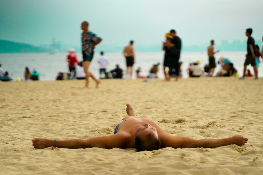 A man lies on the beach while people walk past in the distance