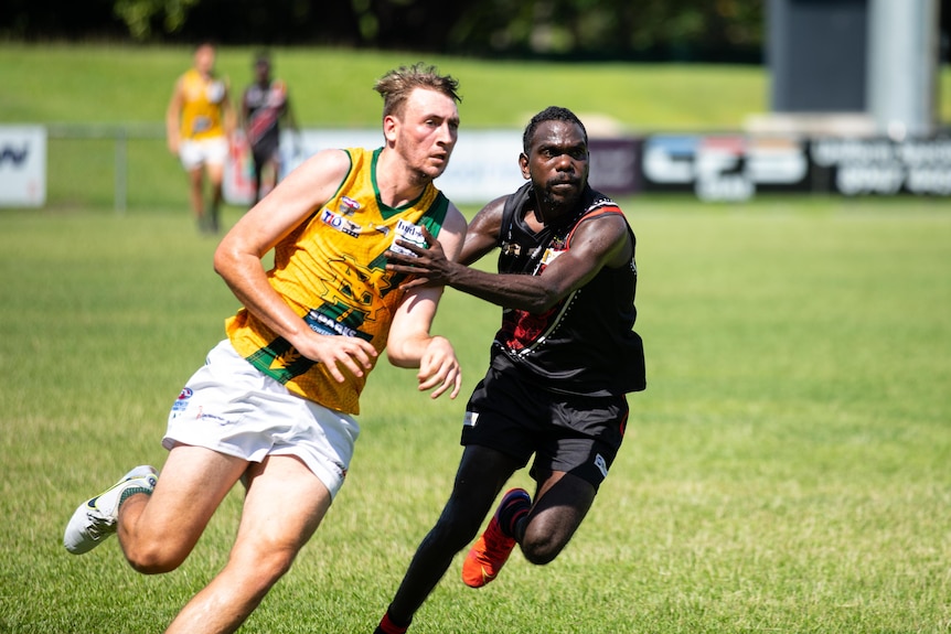 Two men, one wearing a red and black guernsey and another in a yellow and green guernsey, compete in a match of Australian Rules