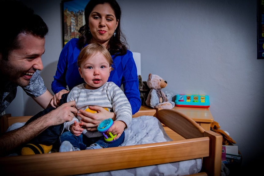 Tom and Nas are smiling as they stand by baby Lachie, who is sitting on a change mat. Lachie is playing with toys.