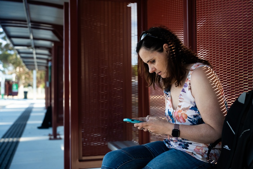 A woman sitting on a bench at a bus station, using her phone.