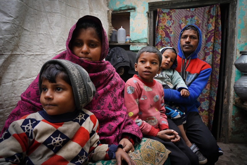 A woman in purple headscarf sits with her three children and a man wearing a blue and red hoodie