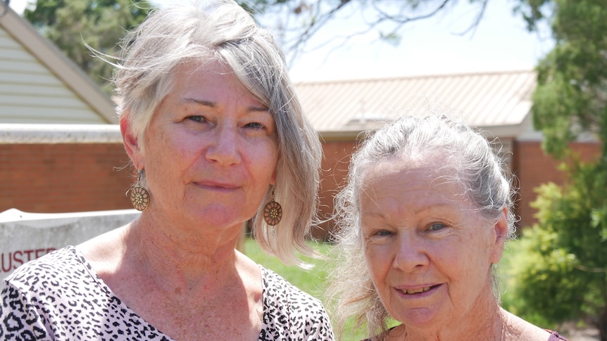 Two elderly women outdoors stand together and smile at the camera. Behind them is a tree and red brick buildings.