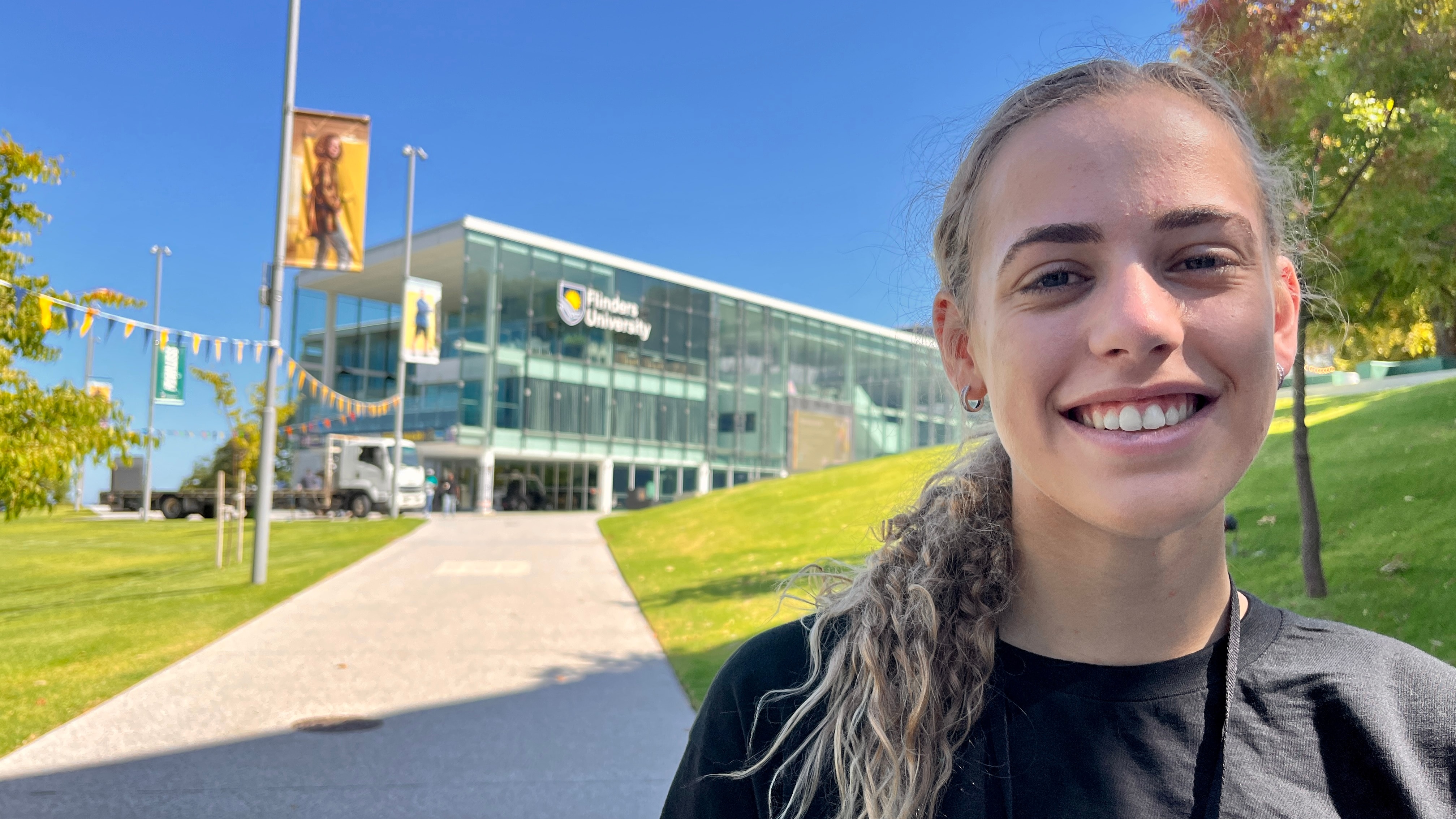 A young woman with a ponytail poses with a smile on a grassed area in front of a glass front university building