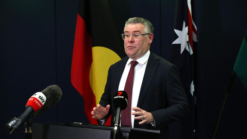 A middle-aged man in a dark suit holds a press conference in front of a lectern. Aboriginal and Australian flags are behind him.