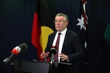 A middle-aged man in a dark suit holds a press conference in front of a lectern. Aboriginal and Australian flags are behind him.