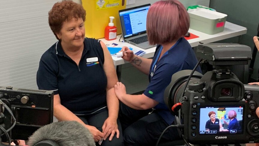 A nurse sitting in front of cameras getting a needle poked into her arm