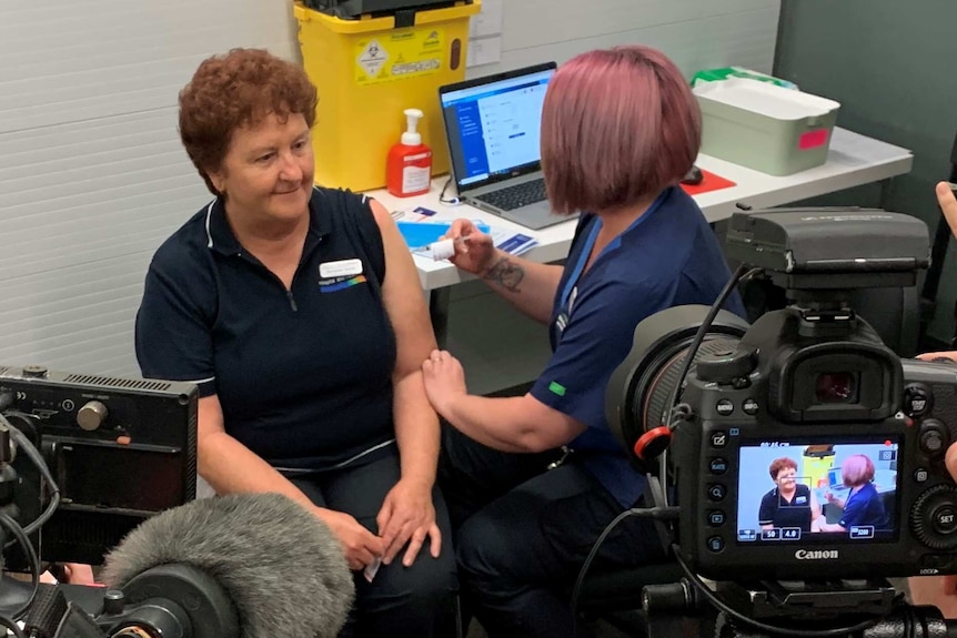 A nurse sitting in front of cameras getting a needle poked into her arm