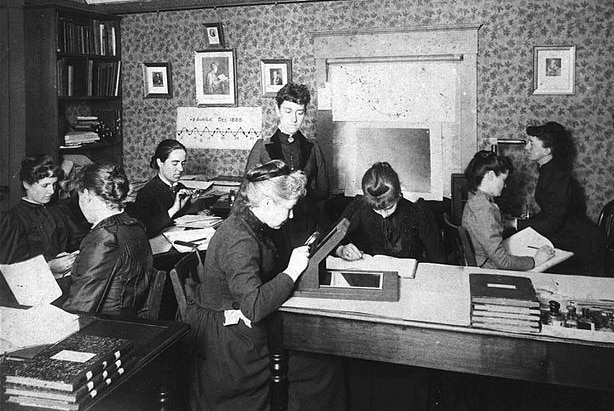 Black and white photo of eight women in a small office looking at photographic glass plates