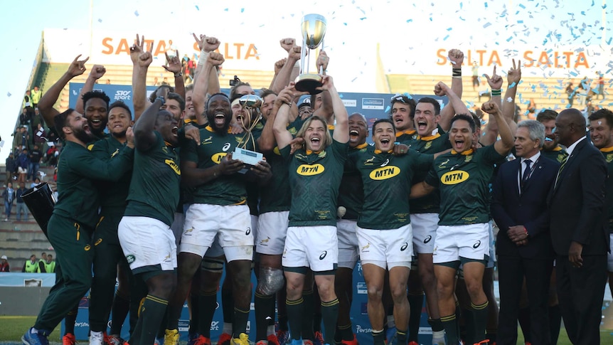A rugby team celebrates on a dais with a trophy as confetti goes off in the background.