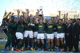 A rugby team celebrates on a dais with a trophy as confetti goes off in the background.