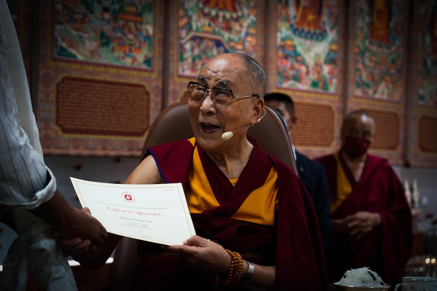 The Dalai Lama's face is lit up with a wide grin, as he hands a piece of paper to a person and shakes their hand