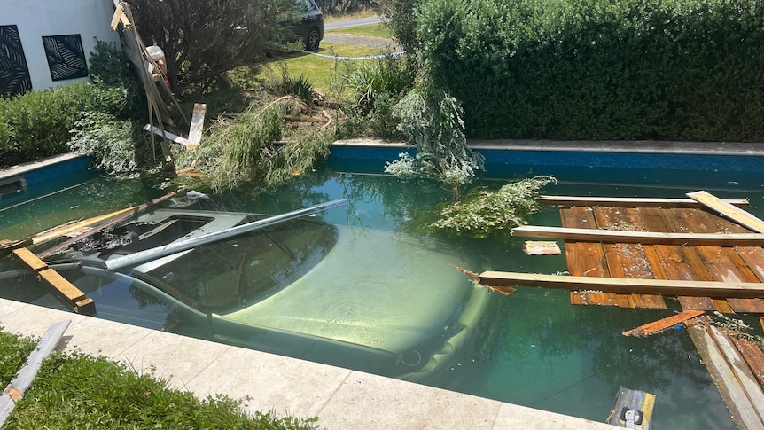 A car is submerged in a backyard swimming pool, with broken fence and debris floating nearby