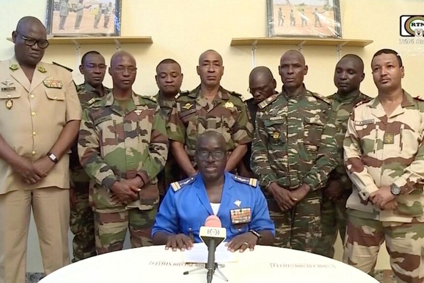 An african man sitting infront of a white table and microphone with nine men in military uniforms behind him