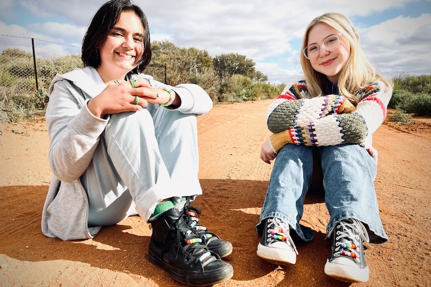 Two teens including a person on the left with medium length black hair and a girl on the right with long blonde hair and glasses