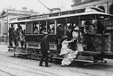 Woman wearing a long dress, hat and shoes, getting onto a Brisbane tram