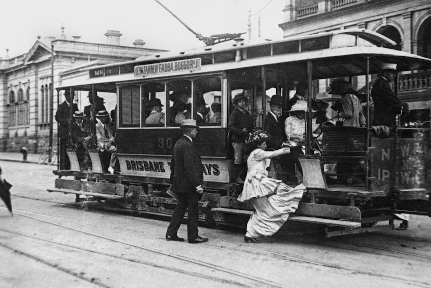 Woman wearing a long dress, hat and shoes, getting onto a Brisbane tram