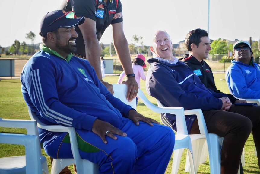 Jamal Mohammed, in a blue cricket uniform, sits on the sidelines of a cricket oval next to other spectators and coaches.