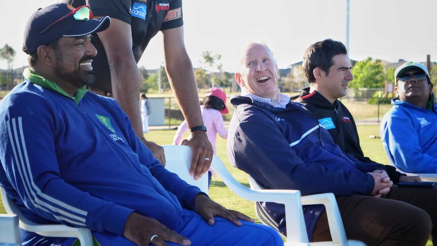 Jamal Mohammed, in a blue cricket uniform, sits on the sidelines of a cricket oval next to other spectators and coaches.