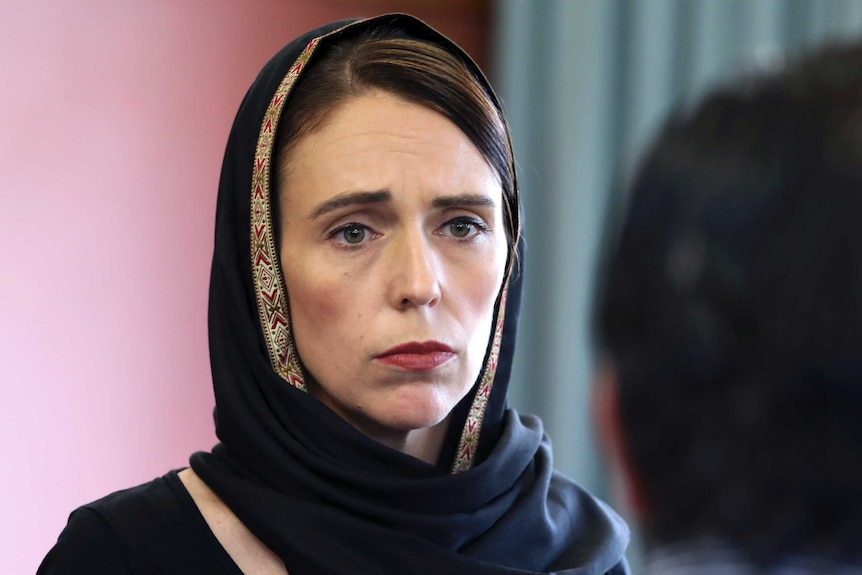 New Zealand Prime Minister Jacinda Ardern looks sad and serious, wearing a black headscarf with gold trim