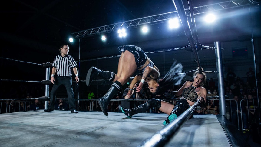 Two female wrestlers in a ring with referee in black-and-white uniform in left corner.