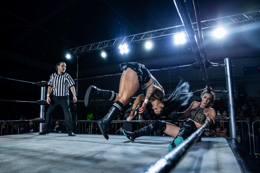 Two female wrestlers in a ring with referee in black-and-white uniform in left corner.