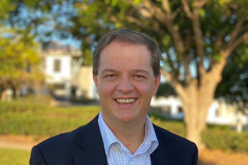 A man in a black jacket stands outside, smiling.