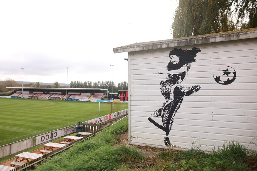 A painting of a woman kicking a ball on the side of a shed overlooking a soccer field
