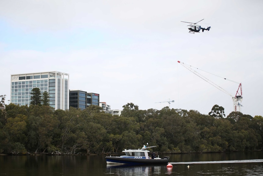 A WA Police helicopter flies over a police boat on the Swan River in Perth with trees and apartment blocks in the background.