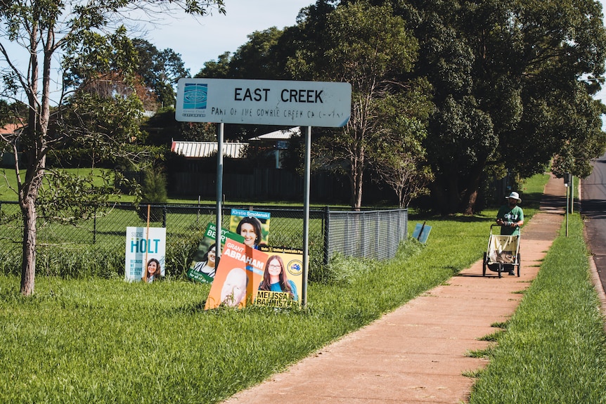 A man pushes a pram next to a roadside park with election corflutes on the side of road.