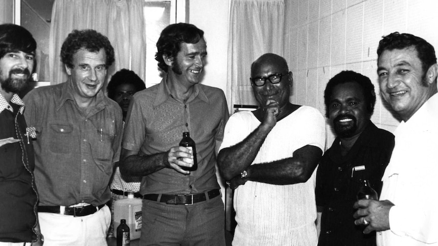 Six jovial men from PNG and Australia stand for a group photo in an office room holding beverages.
