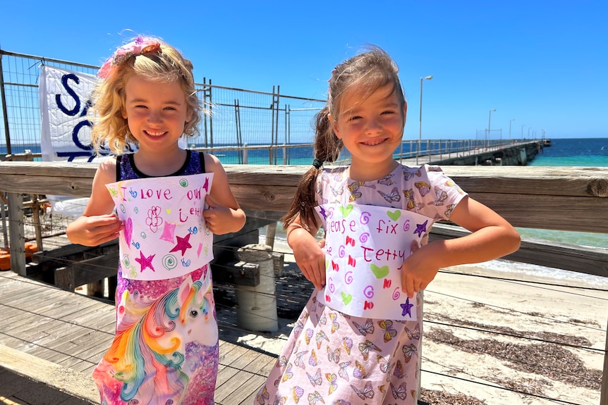 Two litle girls in from of jetty structure holding colourful A4 signs decorated with colourful stars and hearts