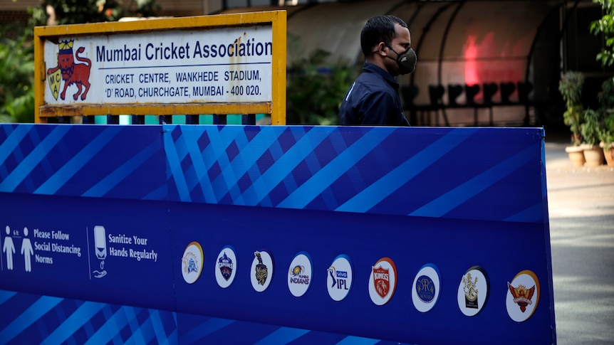 A security guard in a mask stands next to a sign for Wankhede Stadium and the logos of IPL clubs