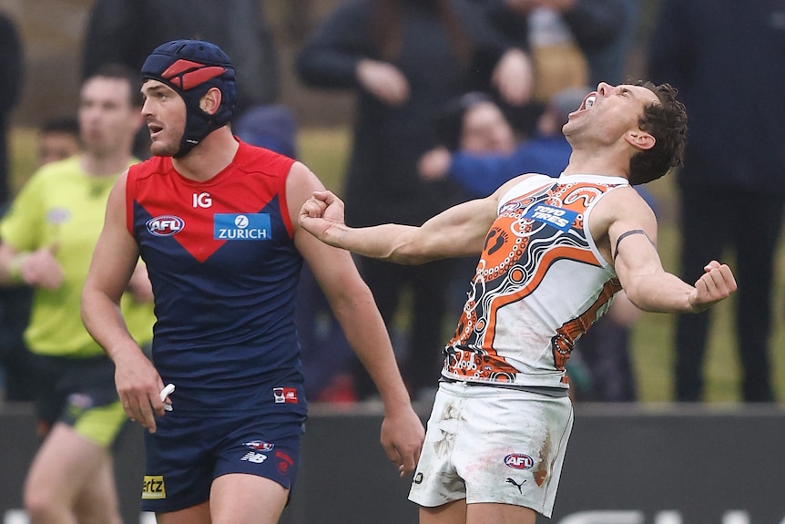 A GWS player closes his eyes, throws his arms wide and roars in celebration after a goal as a dejected defender looks on.