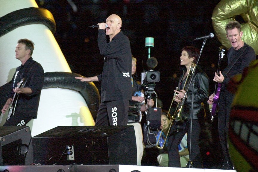 Midnight Oil stand on large stage at Olympics closing ceremony. The word 'Sorry' is on their pant leg