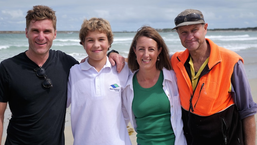 Four people stand on a beach with arms around shoulders smiling