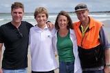 Four people stand on a beach with arms around shoulders smiling