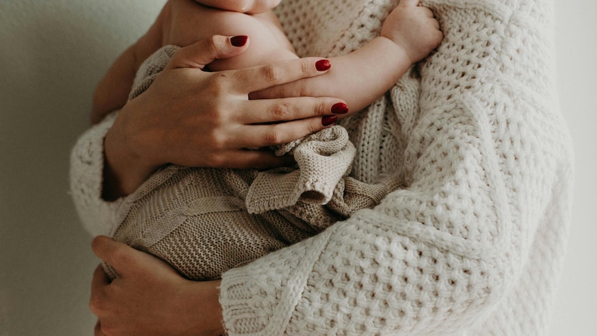 A close up of a woman in beige knitwear holding a baby, their faces are out of frame