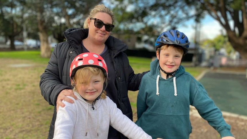 A blonde woman with sunglasses stands with her hands on the shoulders of a young boy and girl wearing bike helmets.