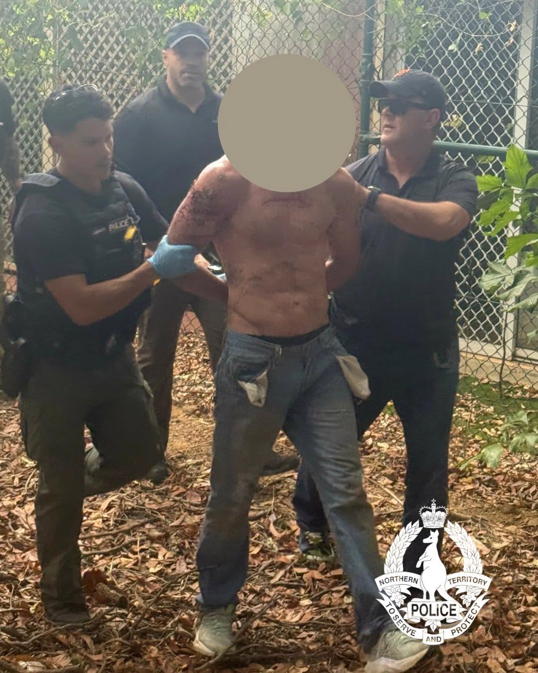 A shirtless man in jeans with his face blurred is surrounded by three police officers who hold his arms