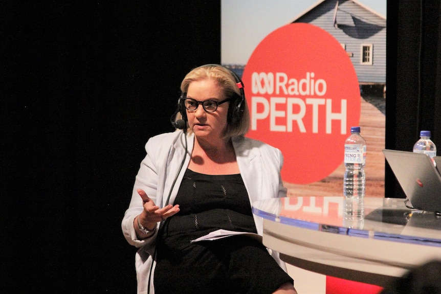 A blonde woman with black glasses sits in front of an ABC Perth banner.