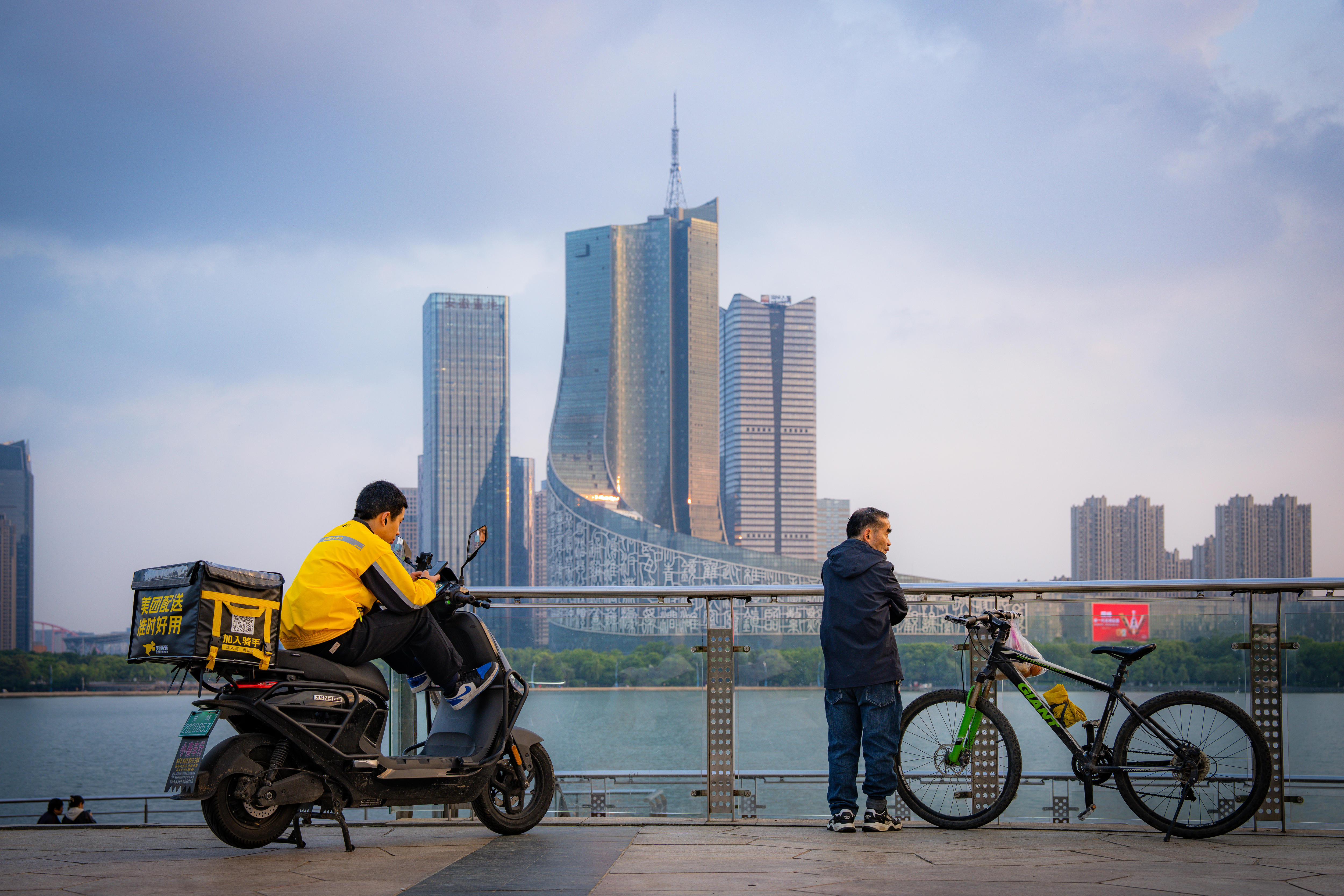 A cityscape with a modern building behind a lake, and a man on a scooter and a man with a bike on the bridge in the foreground