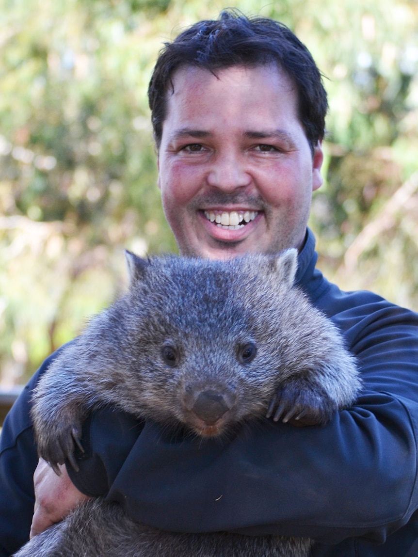 A smiling man holds a wombat in his arms